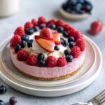 A pink cheesecake topped with raspberries, blueberries, and sliced strawberries on a white plate.