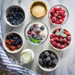 Bowls of fresh and frozen berries, sugar, and cream arranged on a marble surface.