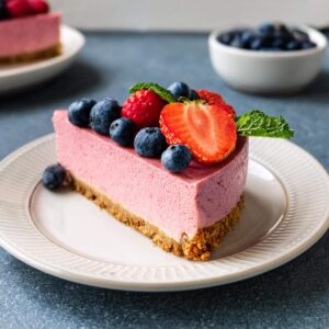 A single slice of pink cheesecake with blueberries, raspberries, strawberries, and mint on a white plate.