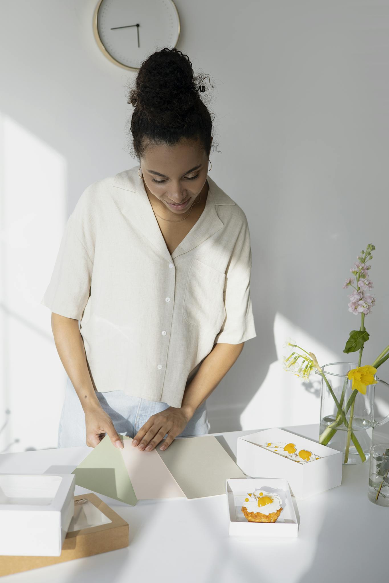 A young woman packages cupcakes in a sunlit modern kitchen with flowers nearby.